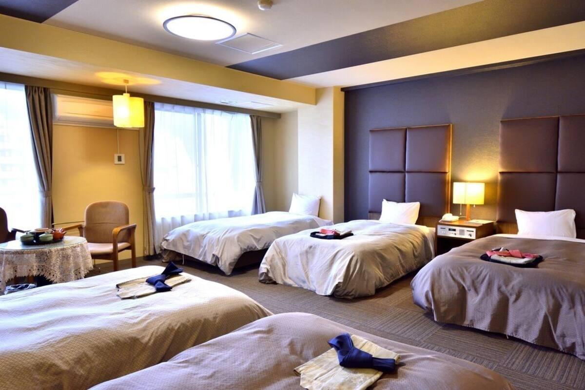 Ein Hotelzimmer mit drei Betten, einem kleinen Tisch mit Lampe und Blick auf die Stadt.