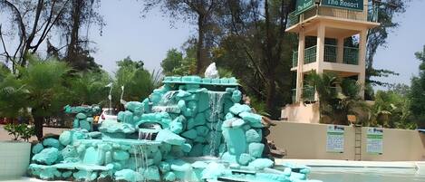A turquoise rock waterfall feature in a resort setting with a viewing tower labeled "Whispering Woods The Resort".