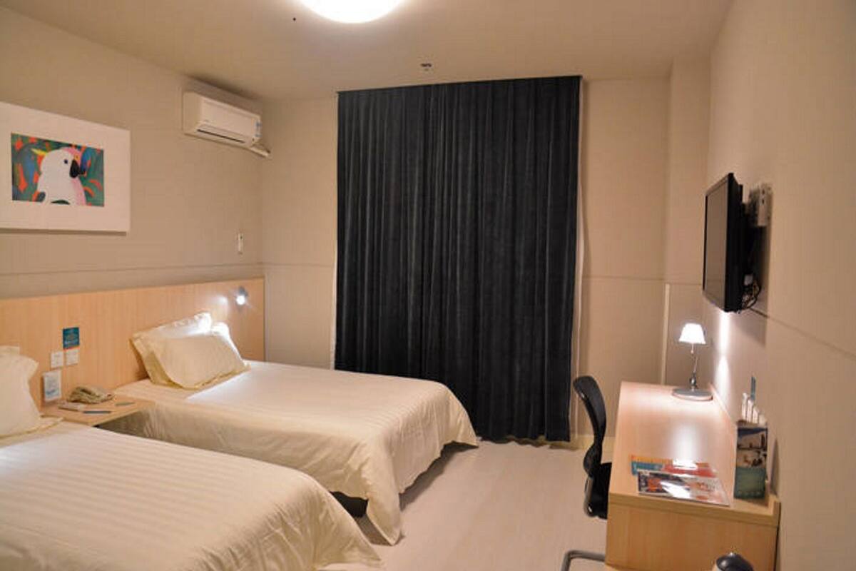 A hotel room with two beds, a desk with a chair, a TV mounted on the wall, and an air conditioning unit.