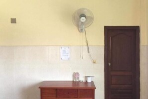 A room with a wooden desk, a wall-mounted fan, a door, and a notice on the wall.