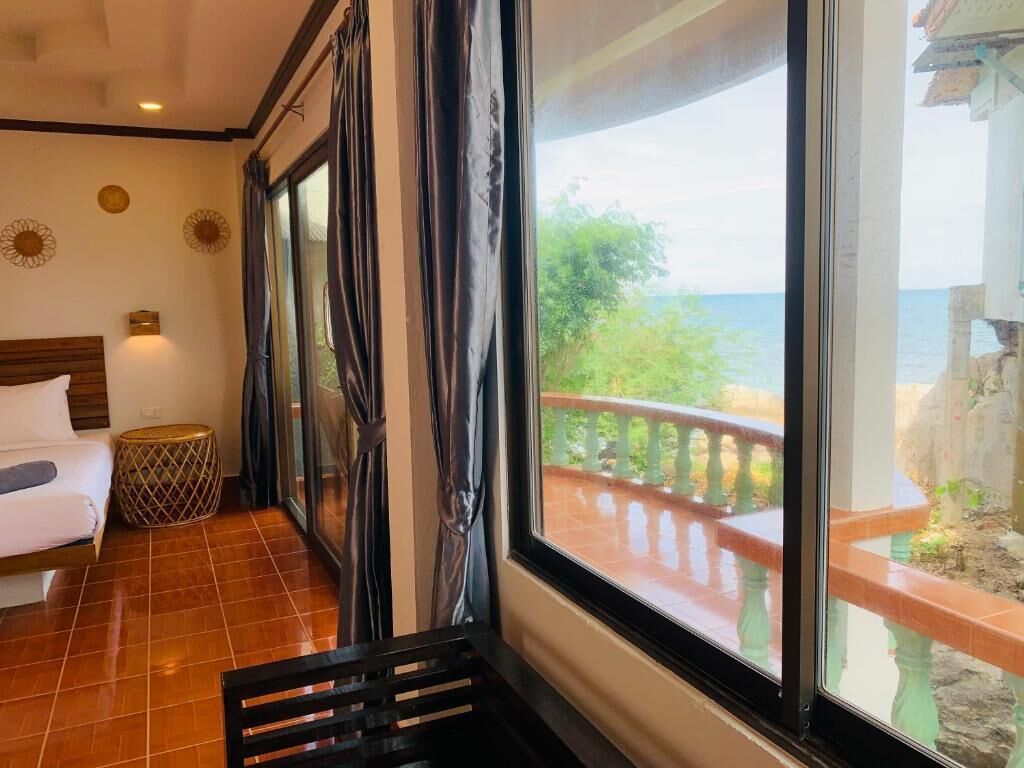 A room with a bed, a small round table, and a view of a balcony with a railing and greenery.