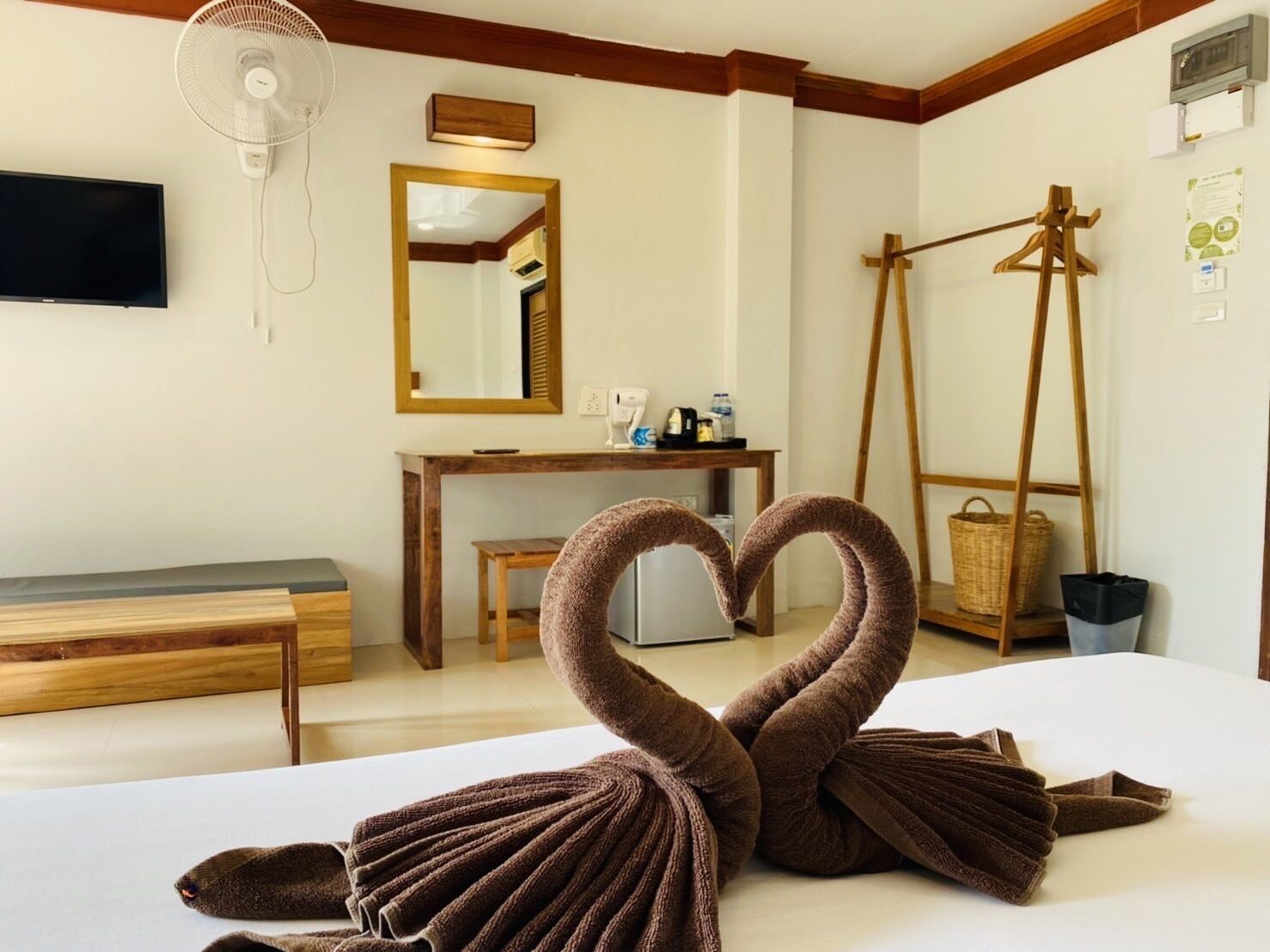 A hotel room with a bed, a wooden bench, a desk, a mirror, a fan, and a wall-mounted television.