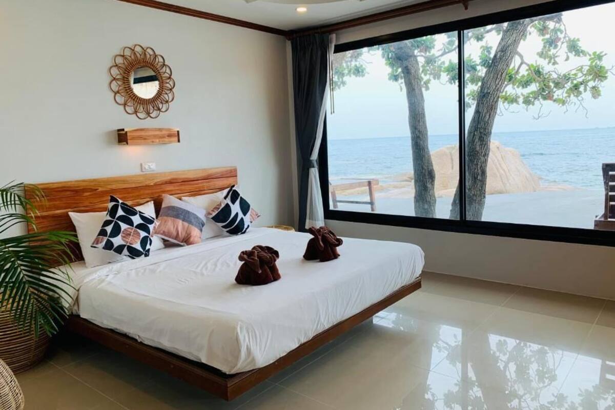 Ruime slaapkamer met een groot bed, een houten hoofdeinde, een decoratieve spiegel en uitzicht op zee door een groot raam.