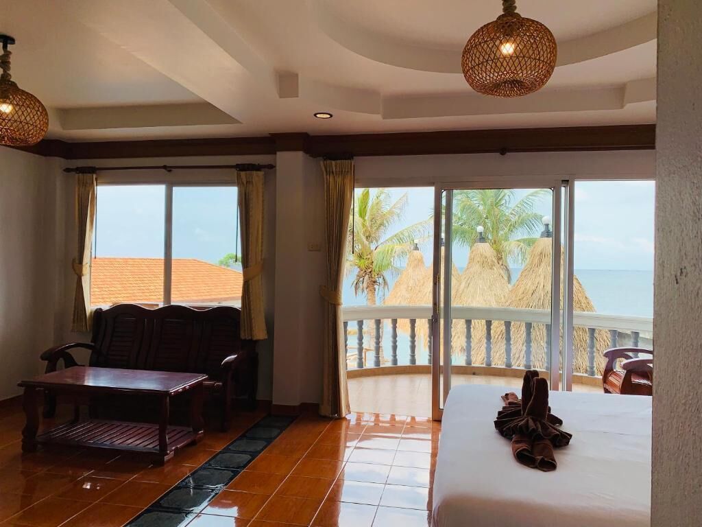 A hotel room with a large bed, a wooden table, and a seating area. It features a balcony with a view of palm trees and the ocean.