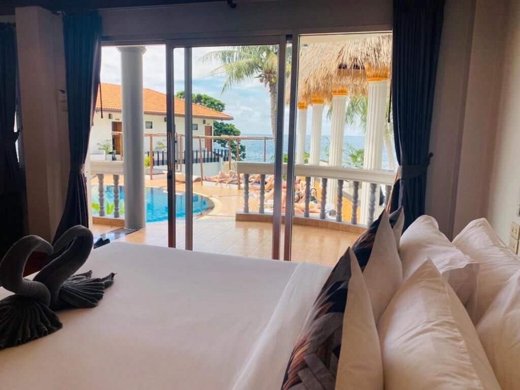A hotel room with a bed, a swan-shaped pillow, and a view of a pool and outdoor area with columns and a thatched roof.