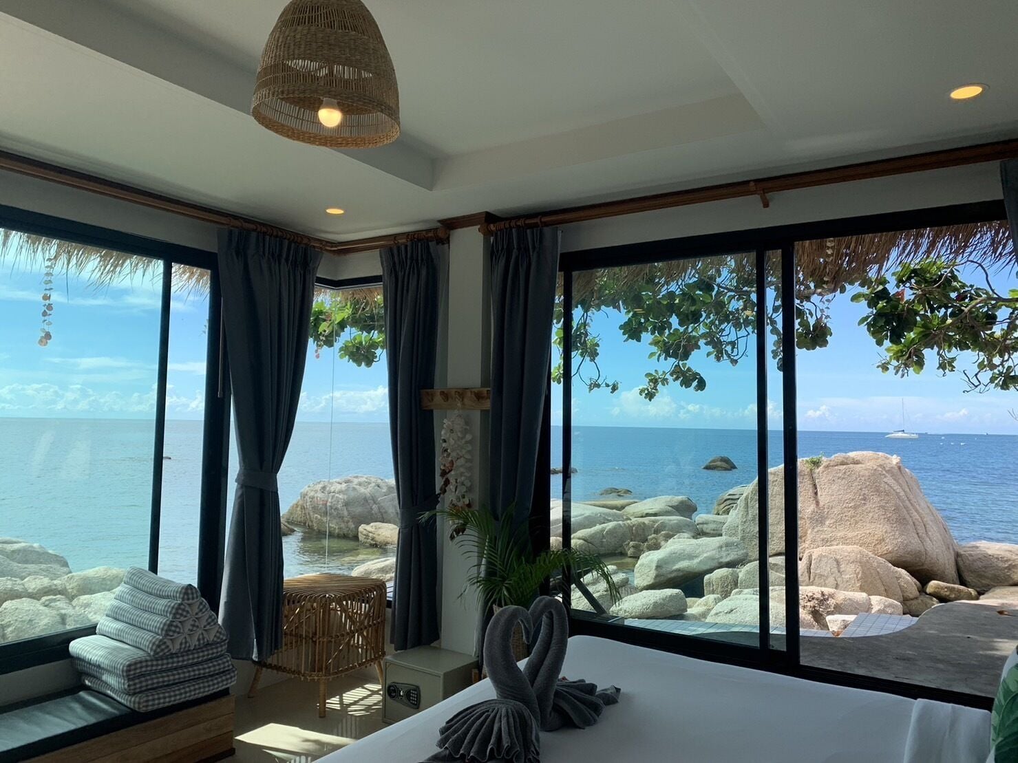 A room with a large window overlooking a rocky coastline and the sea. There is a bed with a white headboard, a wicker chair, and a small table with a plant.
