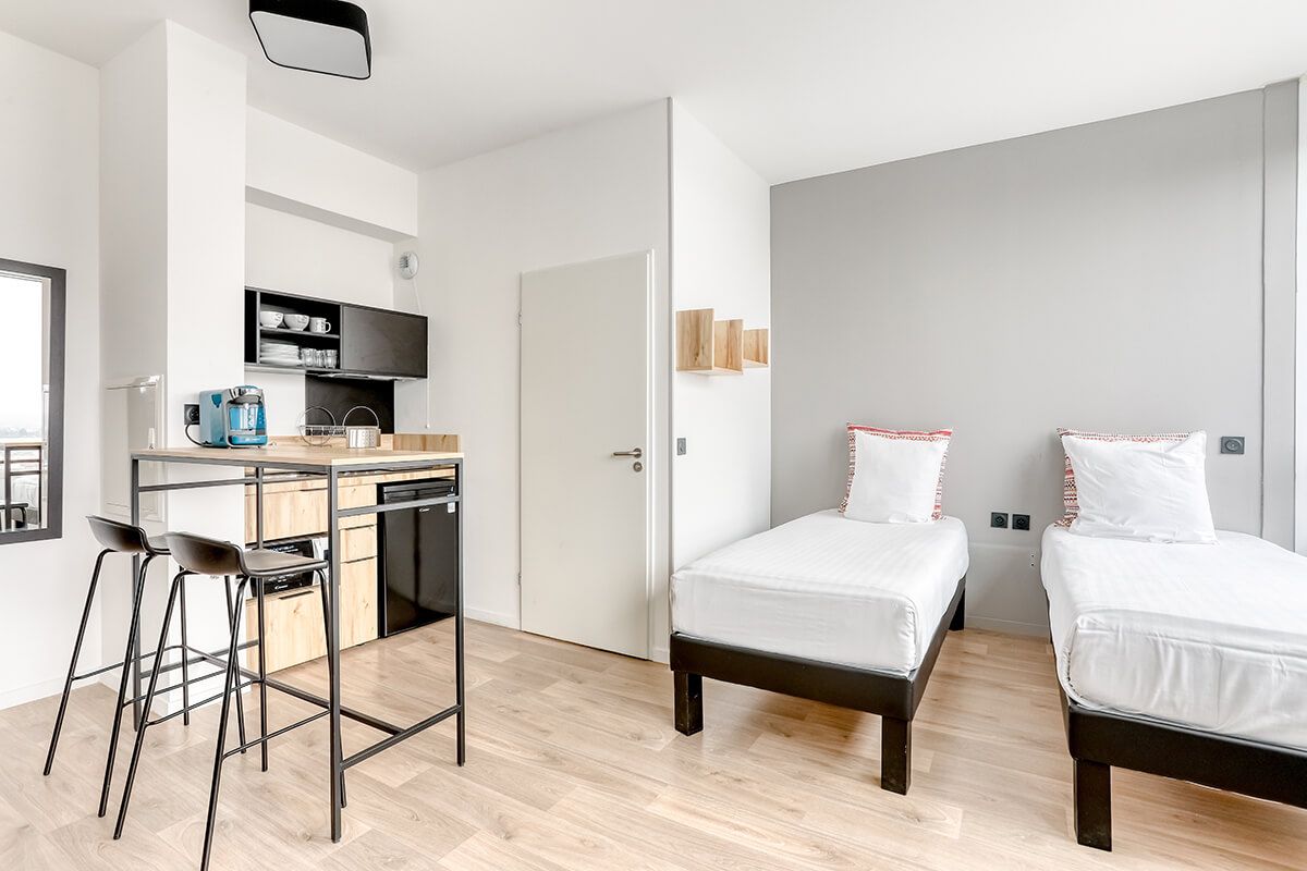 Quarto de hotel moderno com cozinha compacta, duas camas e um banquinho de bar.