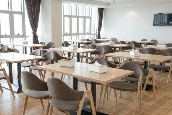 Un espace repas moderne, doté de plusieurs tables et chaises, de grandes fenêtres avec des rideaux et d’un téléviseur à écran plat fixé au mur.