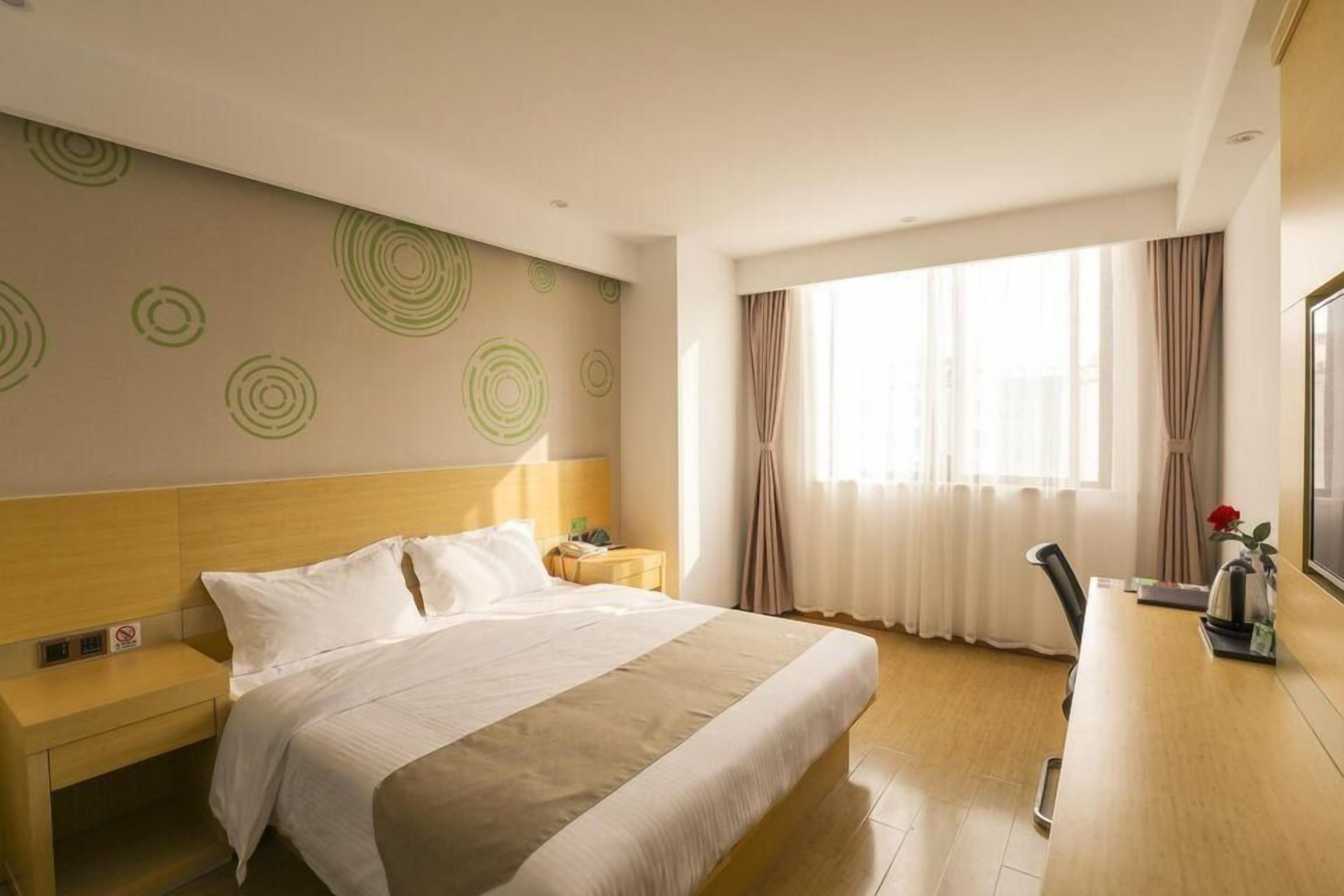 A hotel room with a large bed, a wooden headboard, a bedside table with a lamp, a desk with a chair, and a large window with curtains.