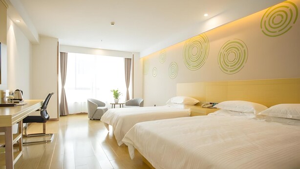 Ein Hotelzimmer mit zwei Betten, einem Schreibtisch mit Stuhl, einem kleinen Tisch mit Pflanze und einem großen Fenster mit Vorhängen.