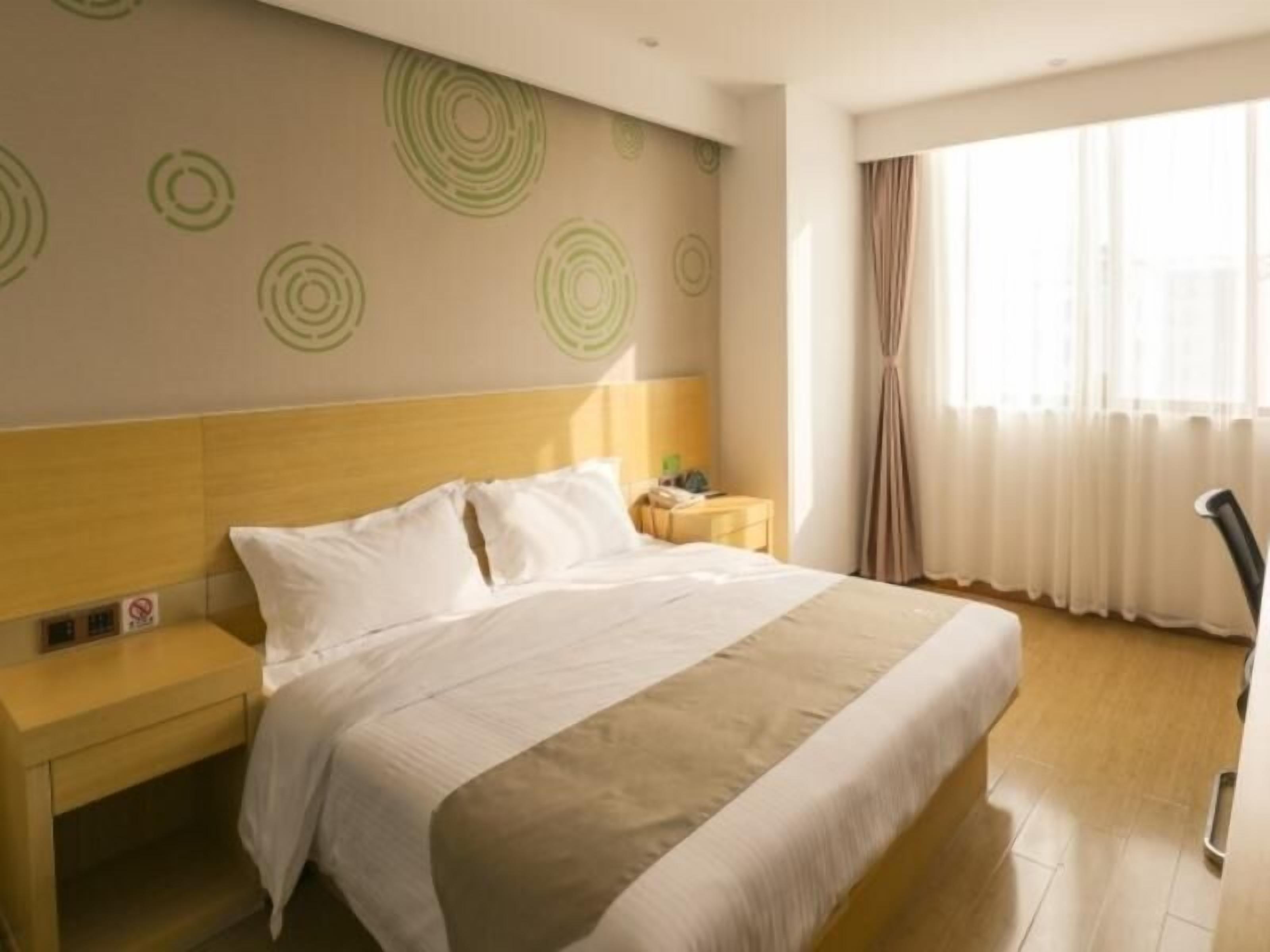 A hotel room with a large bed, a wooden headboard, a bedside table, and a nightstand. There is a window with curtains and a view of the outside.