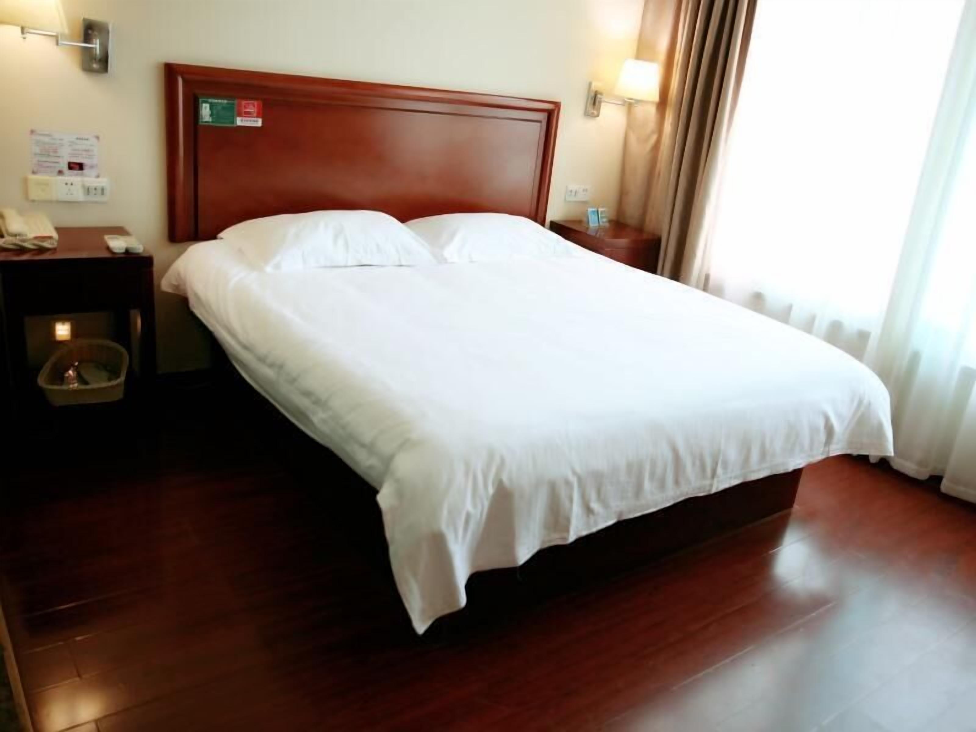 Een hotelkamer met een houten hoofdeinde, een enkel bed met wit beddengoed, een nachtkastje met een lamp, en een bijzettafeltje met een telefoon en een prullenbak.