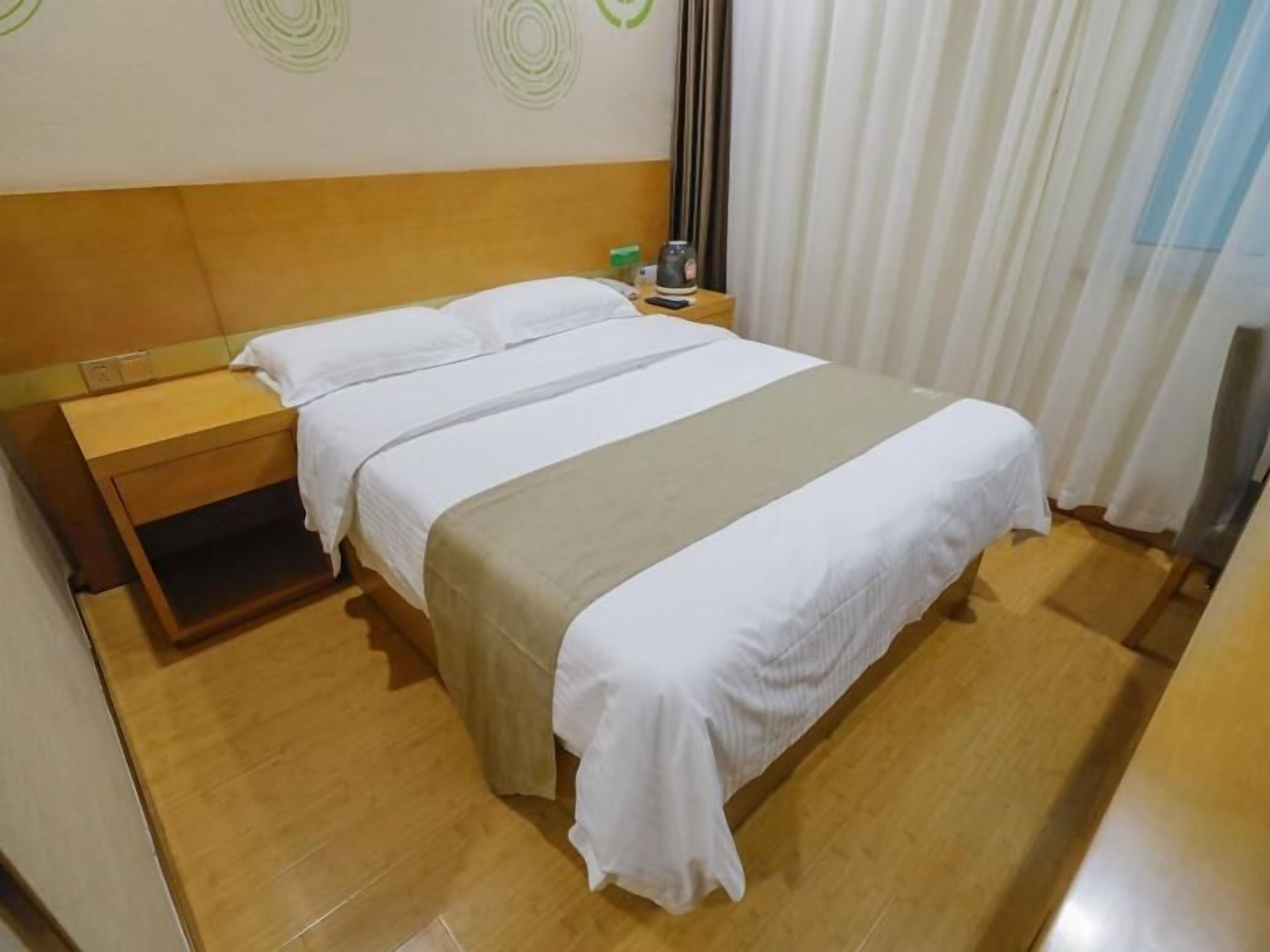 Ein Hotelzimmer mit einem hölzernen Kopfteil, einem Bett mit weißer Bettwäsche, einem Nachttisch mit Lampe und einem Fenster mit Vorhängen.
