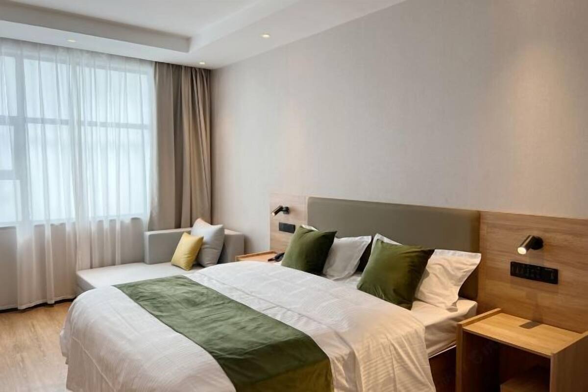 A hotel room with a large bed, a bedside table, a nightstand, and a window with sheer curtains.