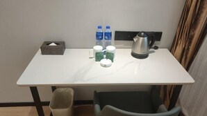 Una habitación de hotel pequeña con una mesa blanca, un cubo de basura, una silla, una caja de pañuelos, dos botellas de agua, una tetera y dos tazas.