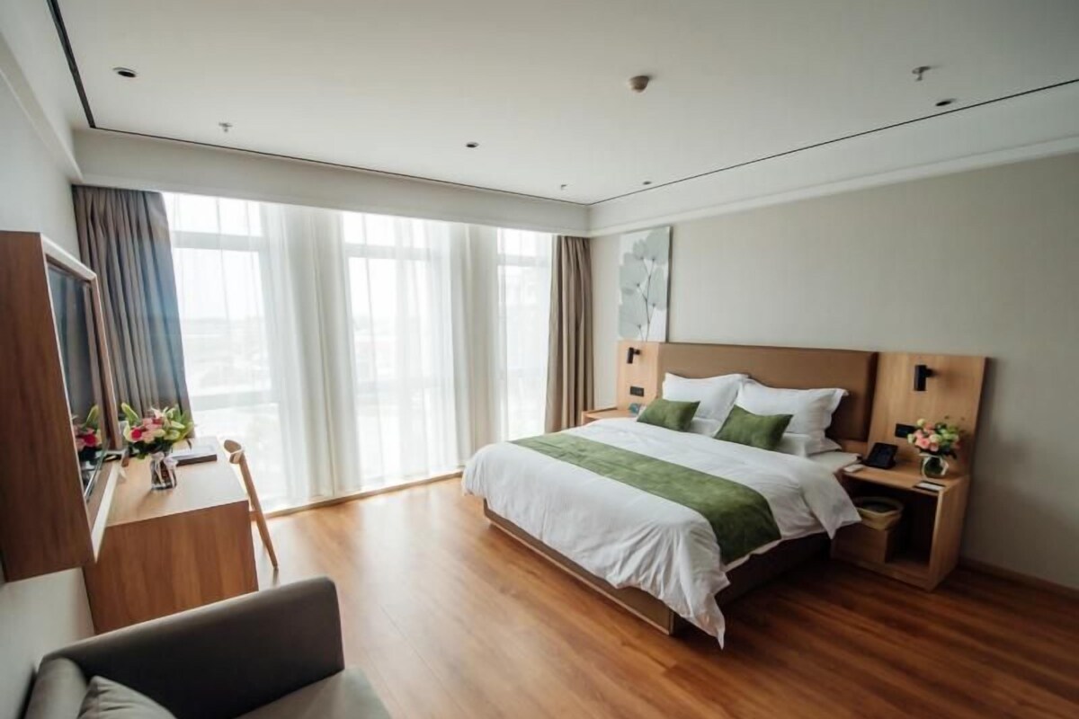 A hotel room with a large bed, a desk with a chair, a bedside table with a lamp, and a large window with curtains.