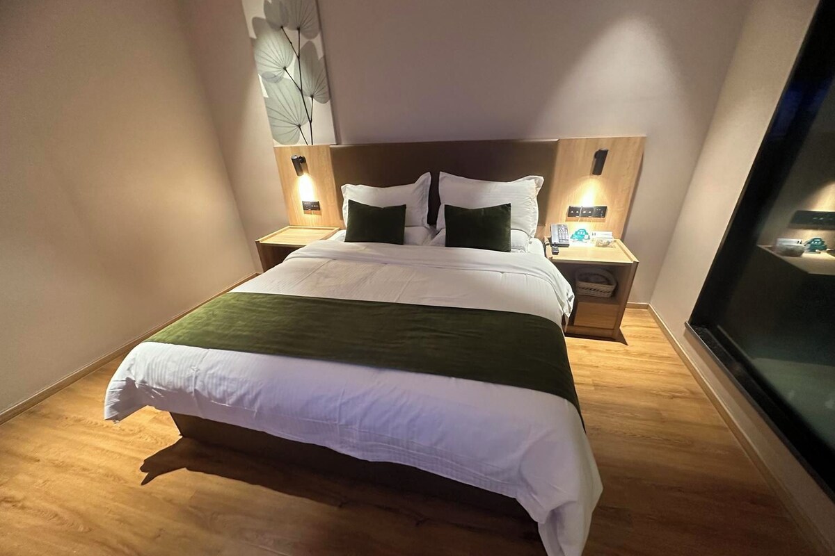Habitación de hotel con una cama grande, dos mesitas de noche, una lámpara de pared y un ventanal.