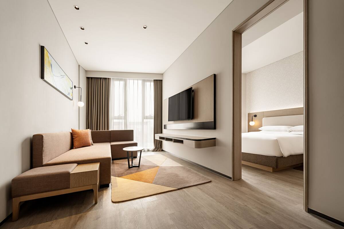 A modern hotel room with a sofa, a small table, a TV mounted on the wall, and a bed with a headboard.