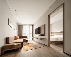 A modern hotel room with a sofa, a small table, a TV mounted on the wall, and a bed with a headboard.
