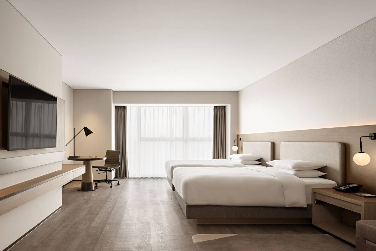 A modern hotel room with a large bed, bedside tables, a desk with a chair, a TV mounted on the wall, and a large window with curtains.