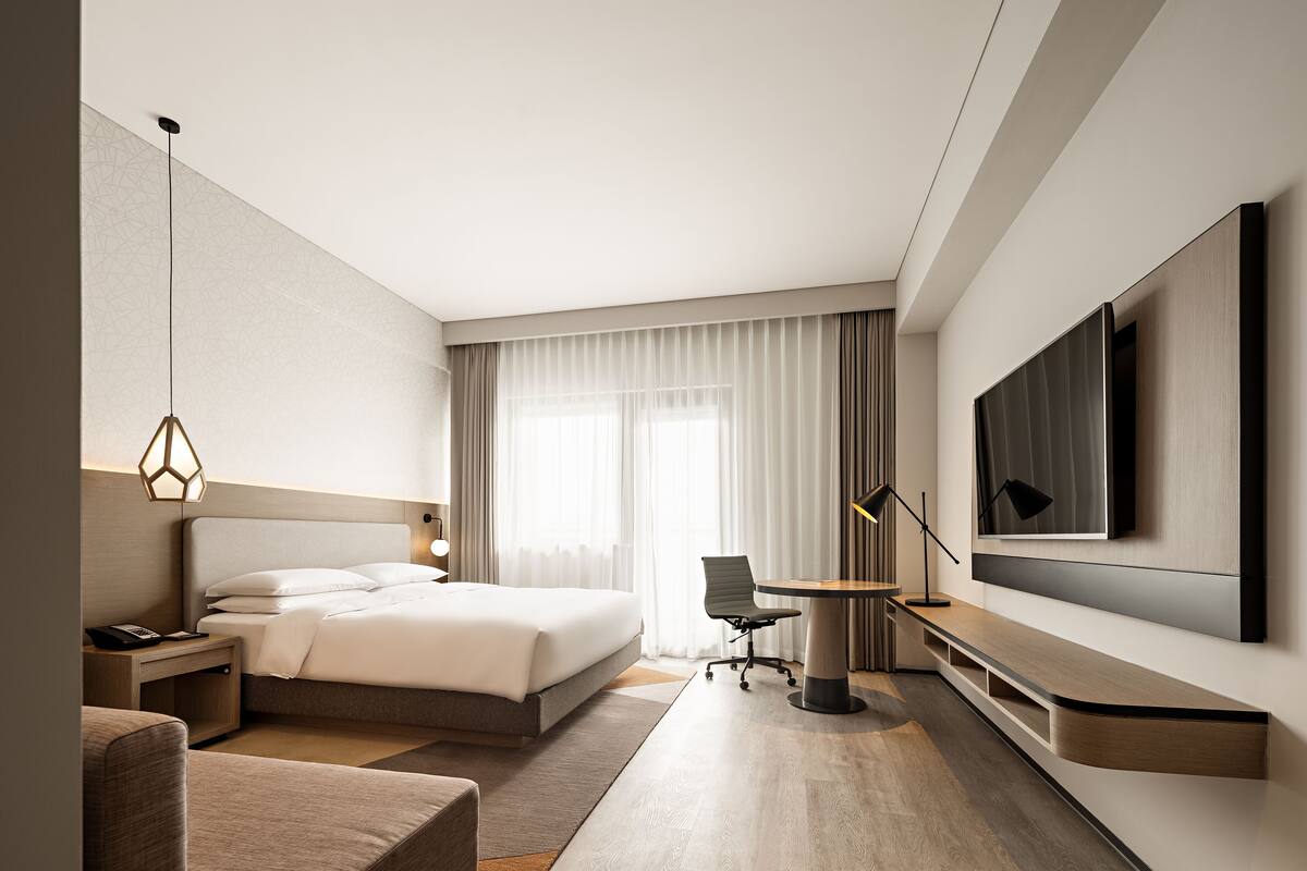 A modern hotel room with a bed, bedside table, lamp, chair, desk, and a large TV mounted on the wall.