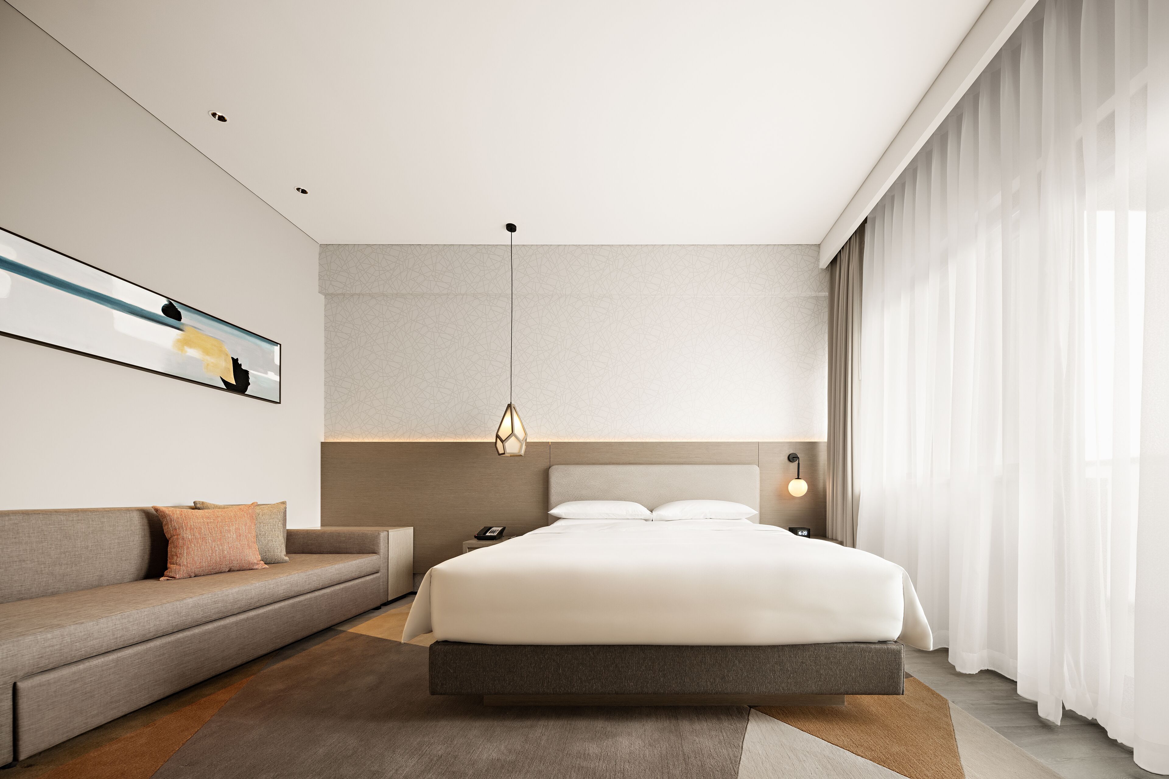 A modern hotel room with a large bed, a sofa, a bedside table, and a wall-mounted artwork.