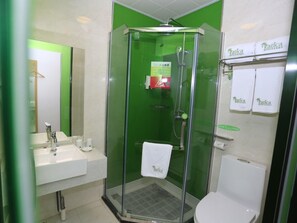 Een badkamer met een douche met glazen wand, een wastafel en een toilet. Handdoeken hangen aan een rek en er is een groen bord aan de muur.