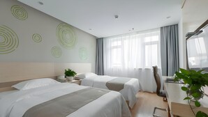 Une chambre d’hôtel avec deux lits, un bureau, une chaise, une plante et une grande fenêtre avec des rideaux.