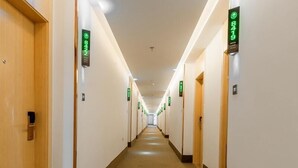 Un couloir d’hôtel avec des portes signalées par des panneaux verts, un sol carrelé et un plafond doté d’un éclairage encastré.