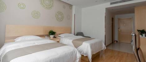 A hotel room with two beds, a wooden headboard, a small bedside table with a plant, and a door leading to another room.