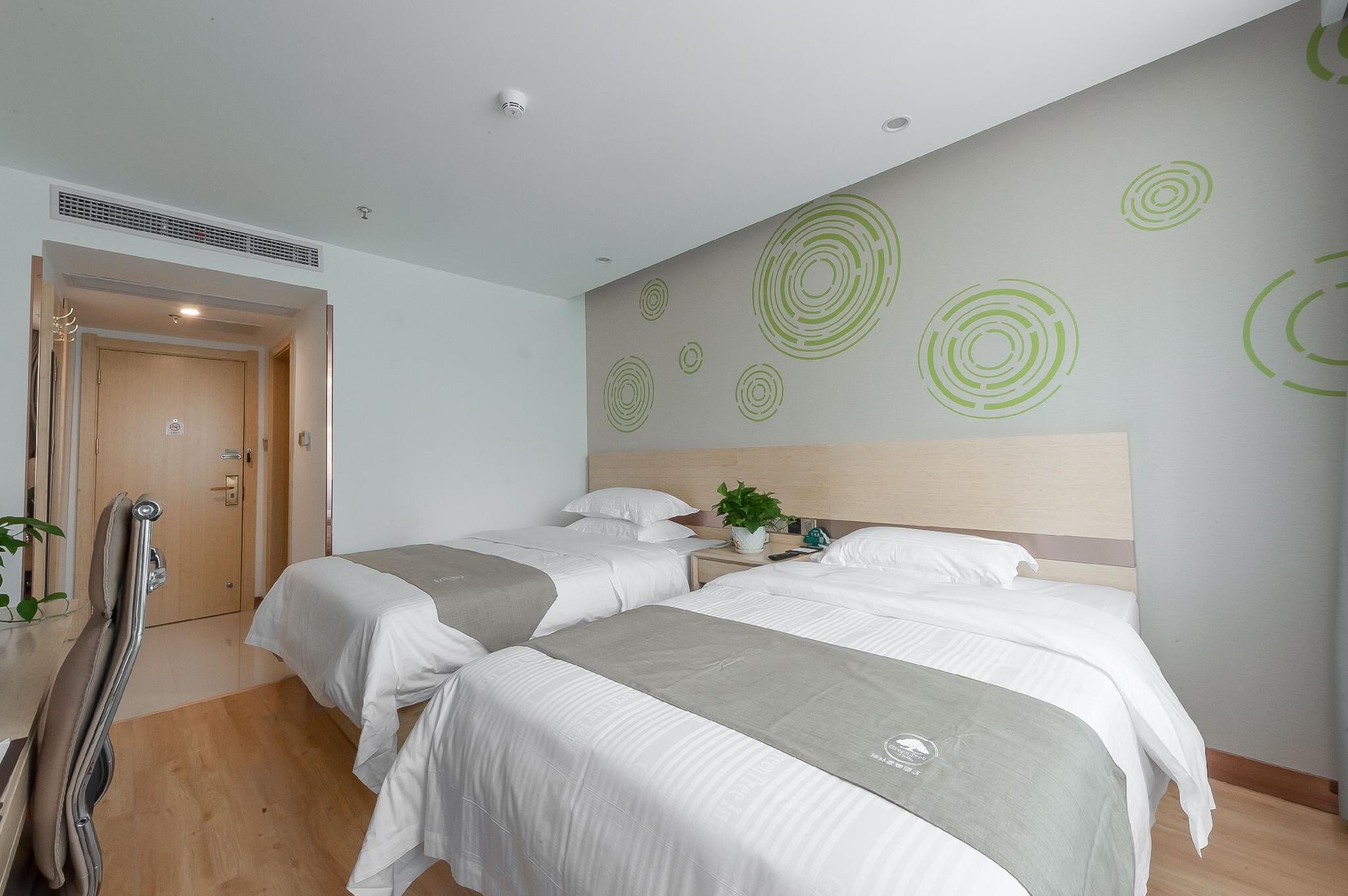 Habitación de hotel con dos camas, cabecera de madera, una planta y una puerta con ventila.