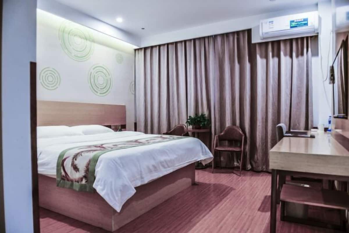 A hotel room with a bed, a desk, a chair, a potted plant, and a large window with curtains.