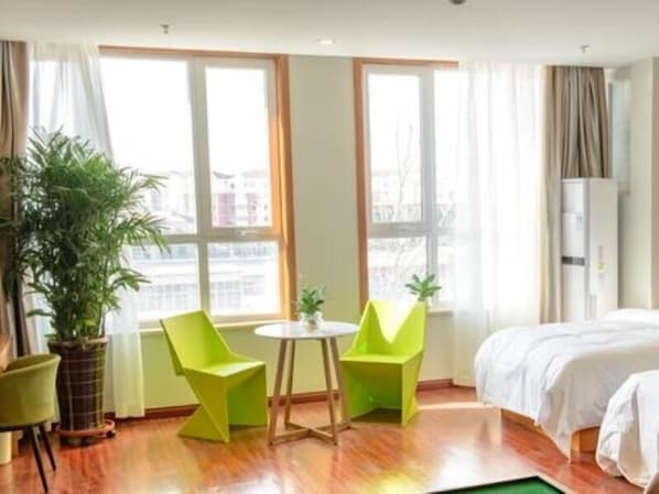 Une chambre d’hôtel avec un lit, deux fauteuils verts, une petite table, une plante en pot et de grandes fenêtres.