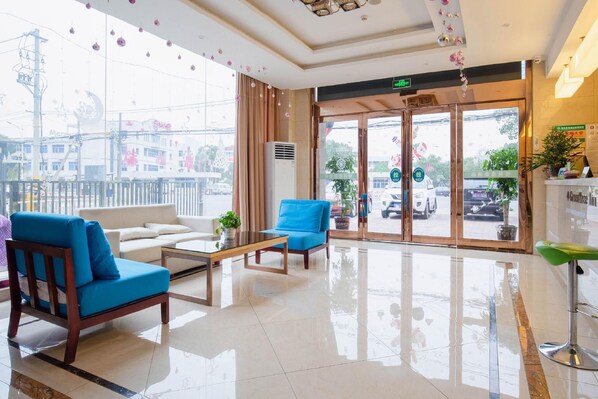 Eine Hotelhalle mit modernem Design, inklusive einer Rezeption, einer Sitzecke mit einer Couch und Sesseln sowie großen Fenstern mit Blick auf die Straße.