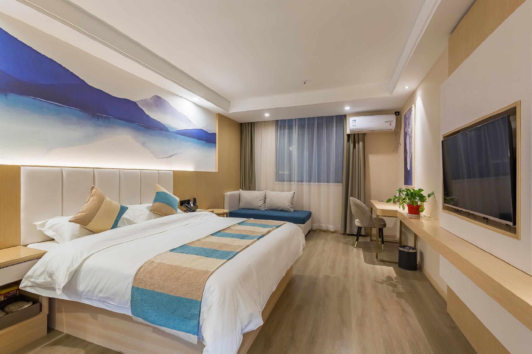 A hotel room with a large bed, a smaller bed, a desk with a chair, an air conditioning unit, a TV, and a painting on the wall.