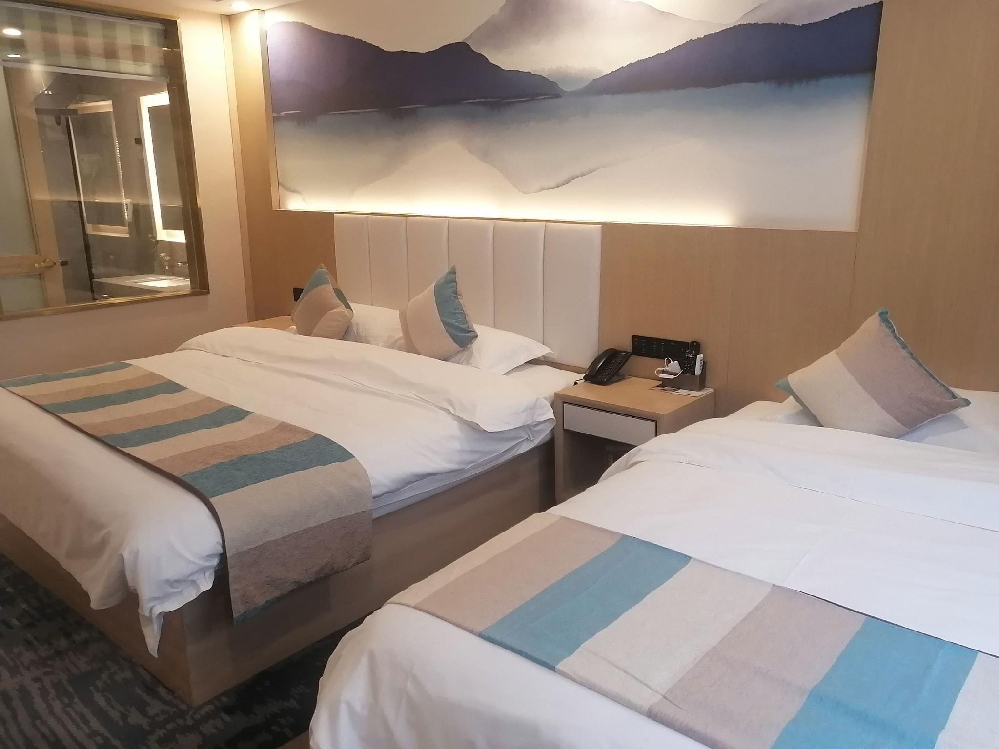 Habitación de hotel con dos camas, una mesita de noche con un teléfono y una lámpara, y un cuadro de paisaje grande en la pared.
