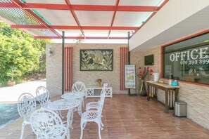 Un espacio al aire libre con sillas y mesas de metal blanco, un cuadro en la pared y una oficina de vidrio con un número de teléfono.