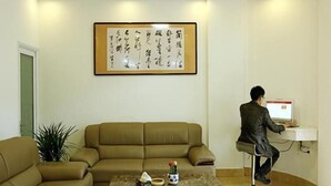 A room with a beige sofa, a coffee table with items, a framed calligraphy piece on the wall, and a person using a computer.