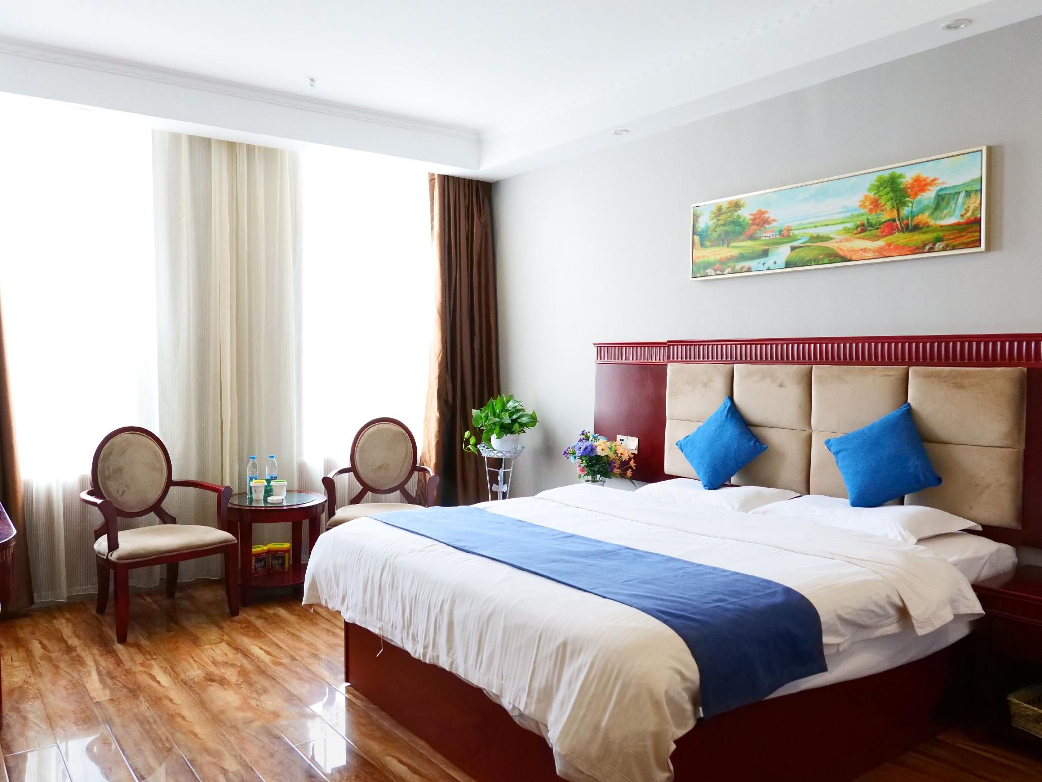 A hotel room with a large bed, a desk with chairs, a window with curtains, and a painting on the wall.
