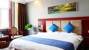 Habitación de hotel con una cama grande, cabecera roja, un cuadro en la pared, una ventana con cortinas y una pequeña mesa con una silla.