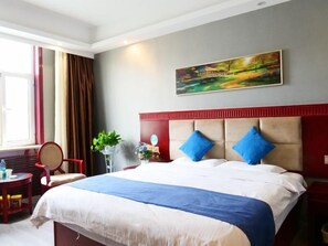 Une chambre d’hôtel avec un grand lit, une tête de lit rouge, un tableau au mur, une fenêtre avec des rideaux et une petite table avec une chaise.