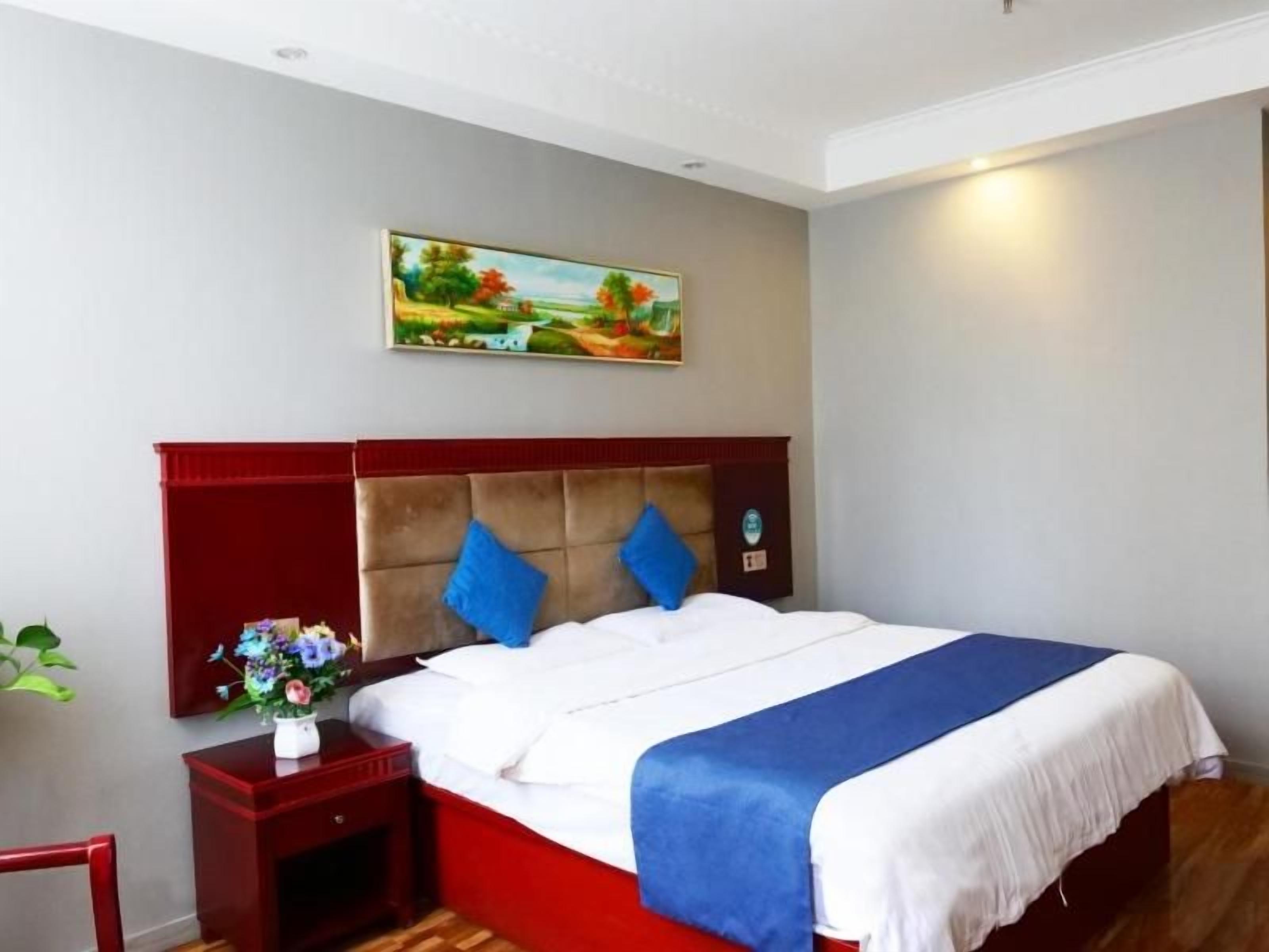 Une chambre d’hôtel avec un lit, une table de chevet, un tableau au mur et une plante.