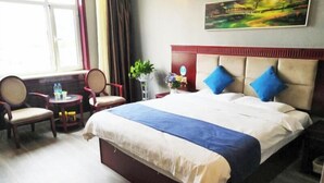 Una cama grande con una colcha azul y blanca, dos almohadas azules y un cabecero rojo. Hay una pequeña mesa con una planta y un jarrón de flores, además de dos sillas. Se ve una ventana con cortinas, y un cuadro cuelga en la pared.