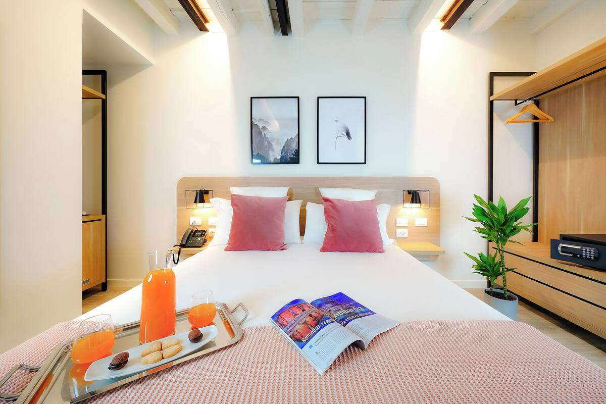 A neatly made bed with pink pillows, a bedside table with a phone, a tray with snacks and orange juice, and two framed pictures on the wall.