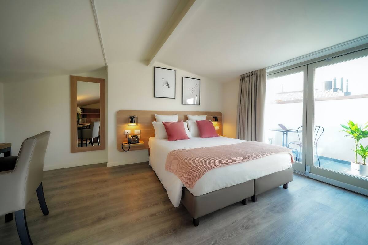 A hotel room with a large bed, bedside tables with lamps, a desk, and a balcony with a plant.