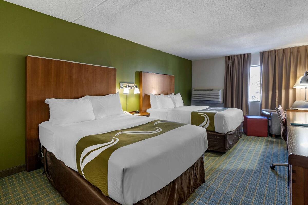 Ein Hotelzimmer mit zwei Betten, einem Schreibtisch, einem Stuhl, einer Lampe und einem Fenster mit Vorhängen.