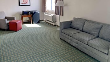 A hotel room with a grey sofa, a red ottoman, a TV on a wooden stand, a lamp, and a window with curtains.