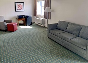 A hotel room with a grey sofa, a red ottoman, a TV on a wooden stand, a lamp, and a window with curtains. - Quality Inn Lexington -horse Park (Lexington)