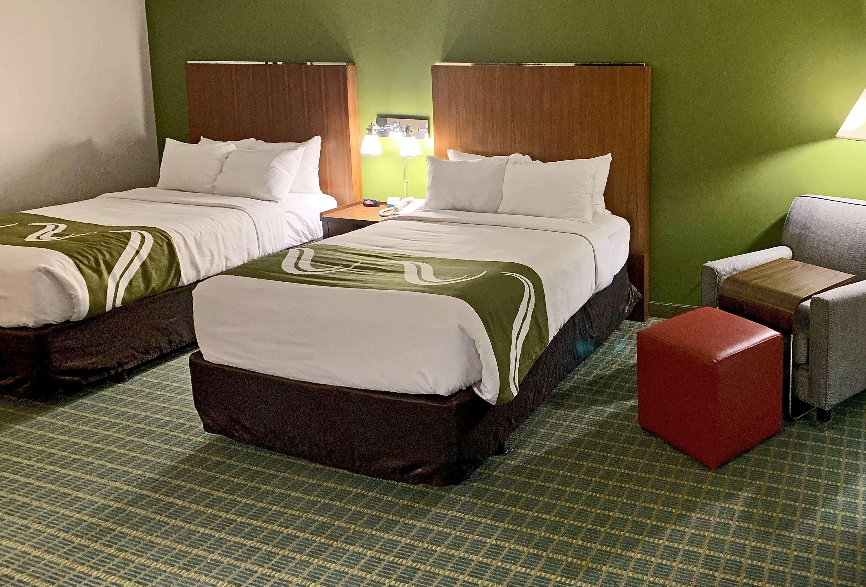 A hotel room with two beds, a red ottoman, and a chair.