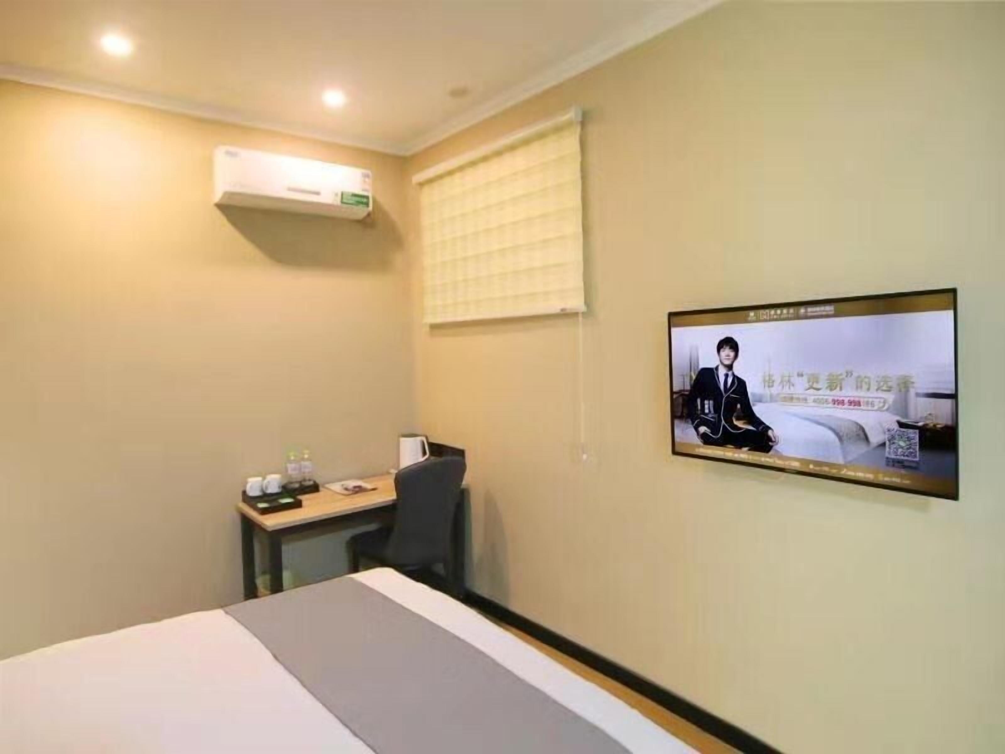 A hotel room with a bed, a desk with a chair, a television mounted on the wall, and an air conditioning unit.